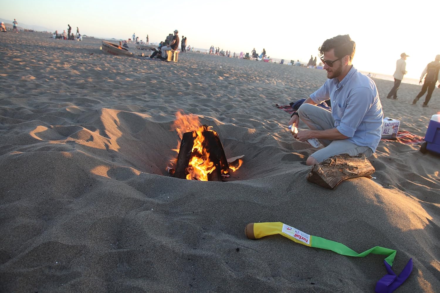A man sits on sand by a fire pit at sunset, with a log, a colorful toy axe, The Original Foxtail Sport perfect for outdoor play nearby, and other beachgoers enjoying the scene in the background.